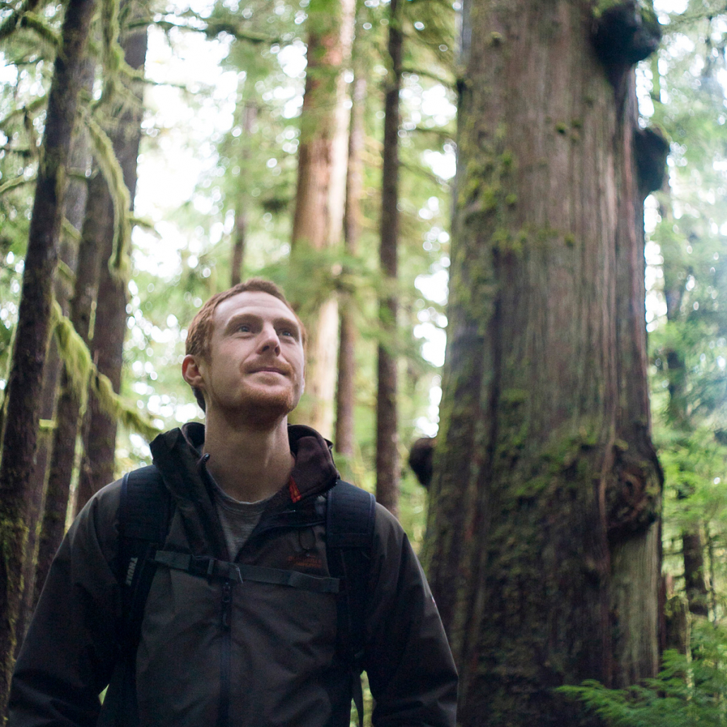 TJ stands in the forefront wearing a black hoodie. Behind him stands a number of old-growth cedars and other ancient trees.