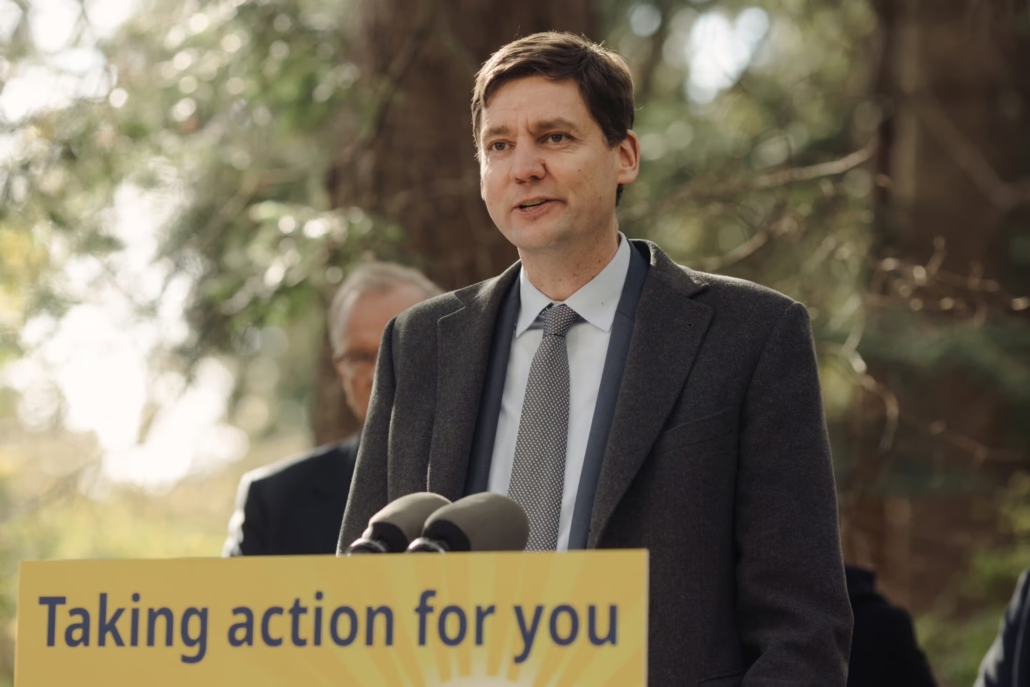 Premier David Eby stands at a yellow podium that reads, "Taking action for you," with trees in the background.